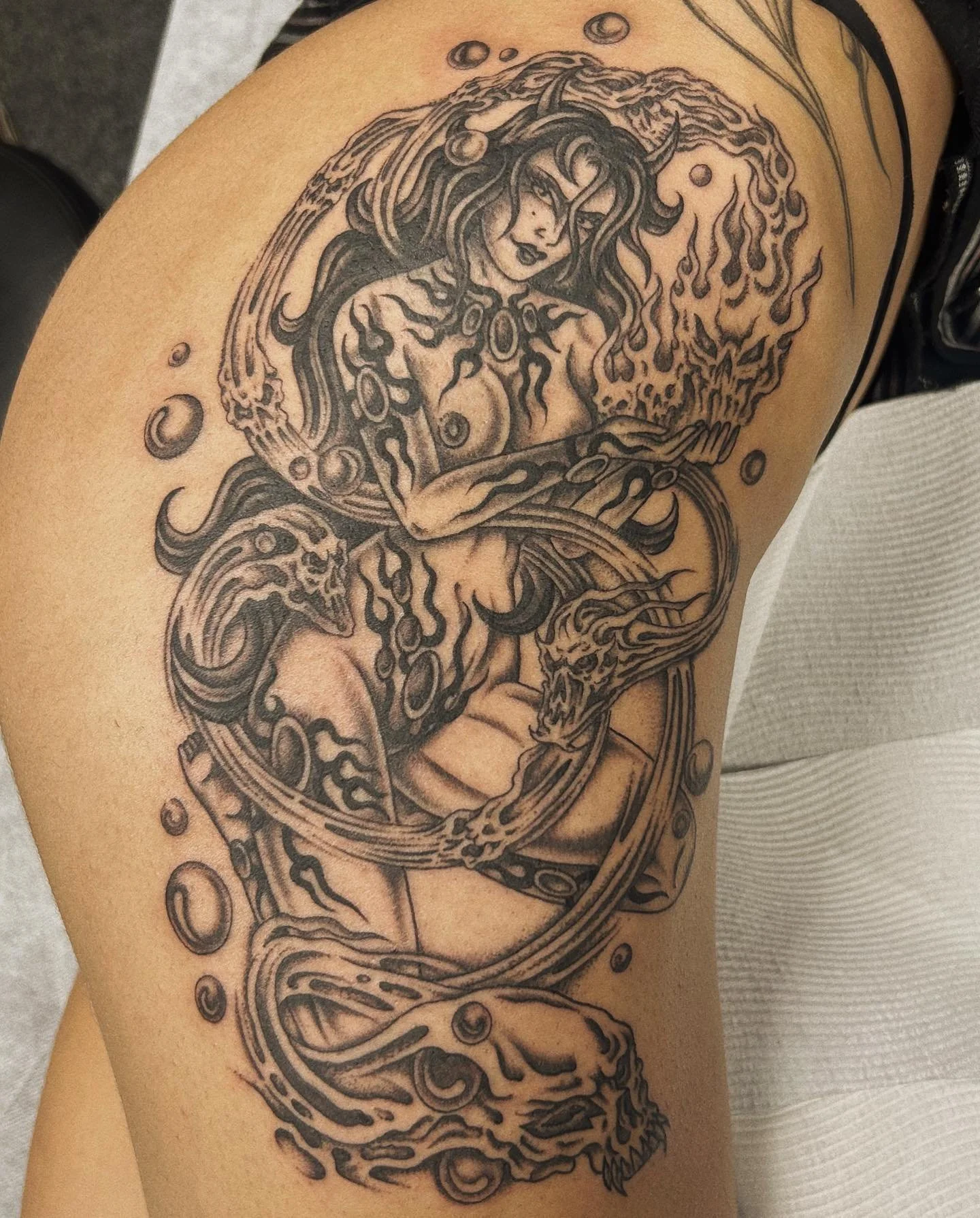 A detailed black and gray tattoo of a mythical creature with a woman's face, dragon-like head, and serpentine body, surrounded by bubbles and abstract flames.