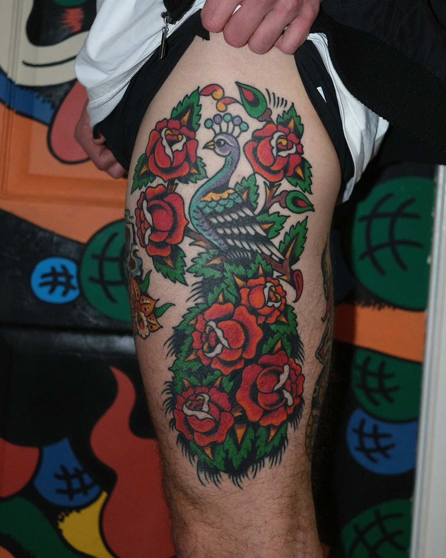 Tattoo of a peacock surrounded by red roses and green leaves on a person's thigh.