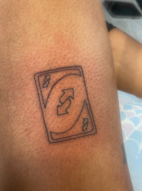 Tattoo of a playing card with a curved arrow, two spade symbols, and two curved lines.