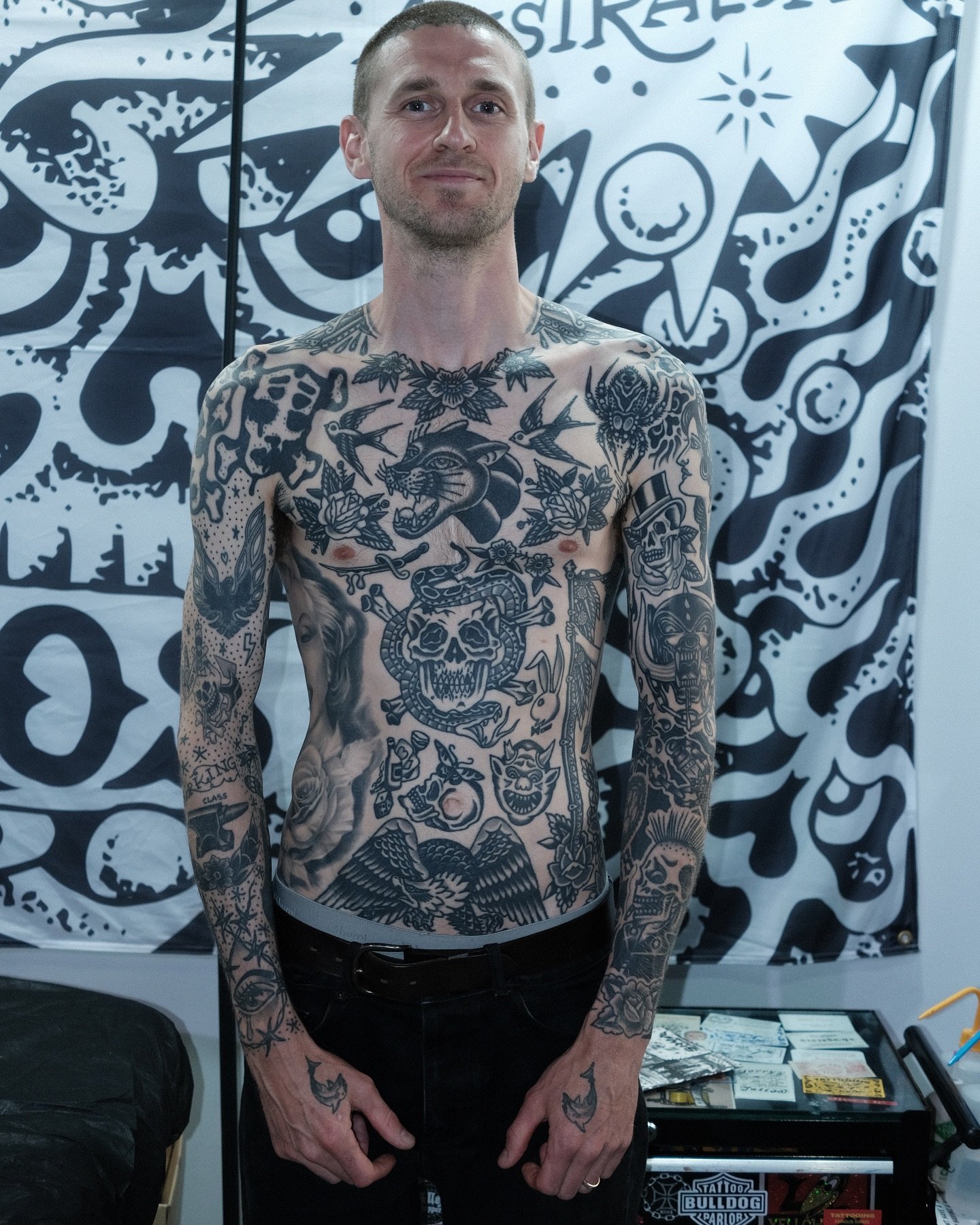 A man with numerous tattoos on his chest and arms standing indoors with a black and white tattoo-themed wall behind him. He has short hair, is shirtless, and is smiling slightly.