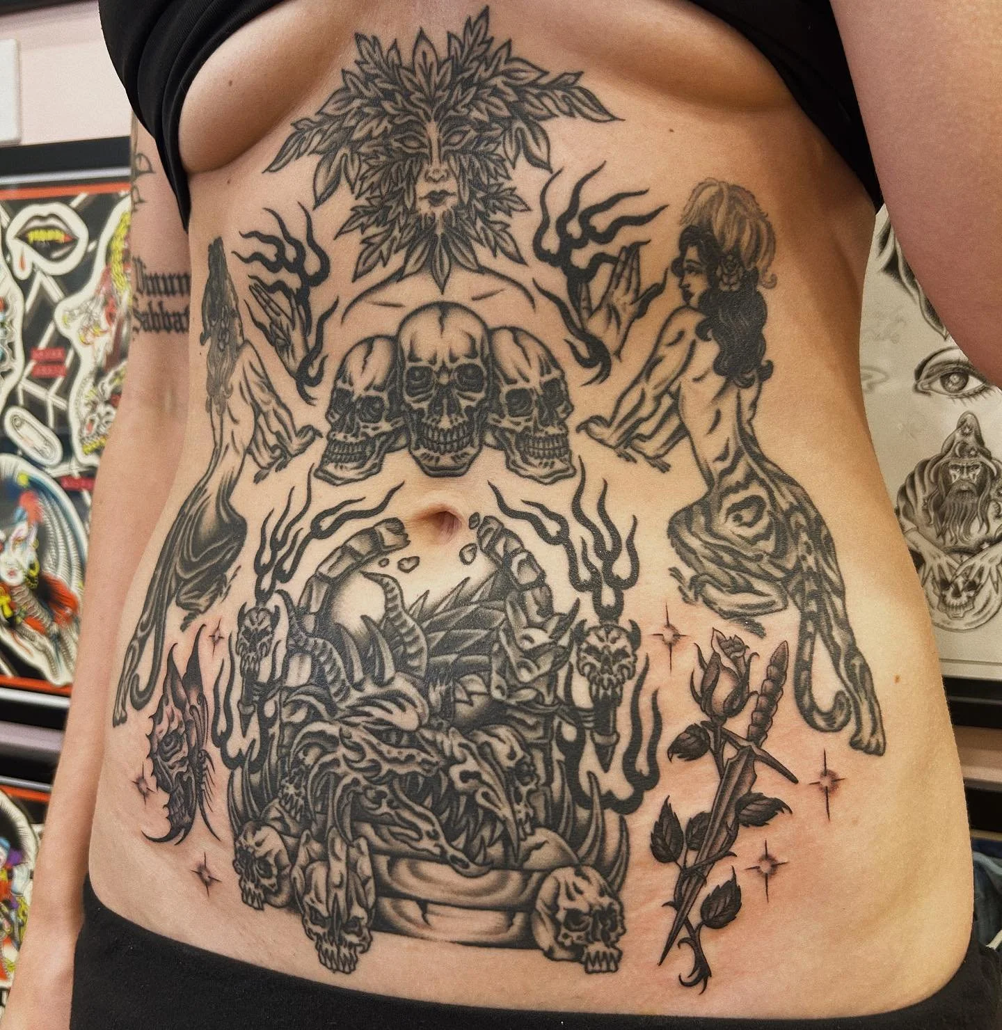 A person's abdomen and side with a large black tattoo featuring skulls, a woman, a lion's head made of leaves, and various mythical and dark imagery, including a scorpion, a dagger with roses, and smoke-like designs.