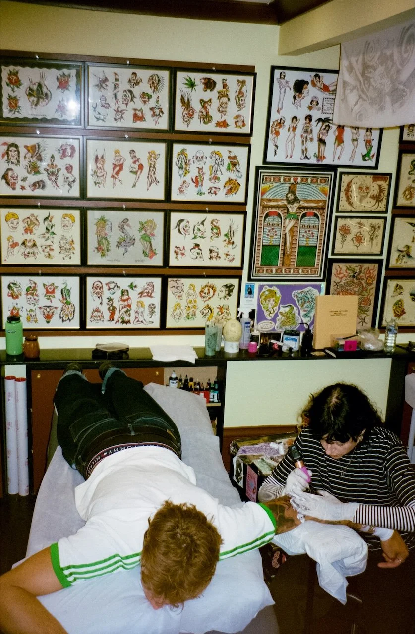 A person gets a tattoo on their arm by a tattoo artist in a tattoo studio with artwork and flash sheets on the wall.