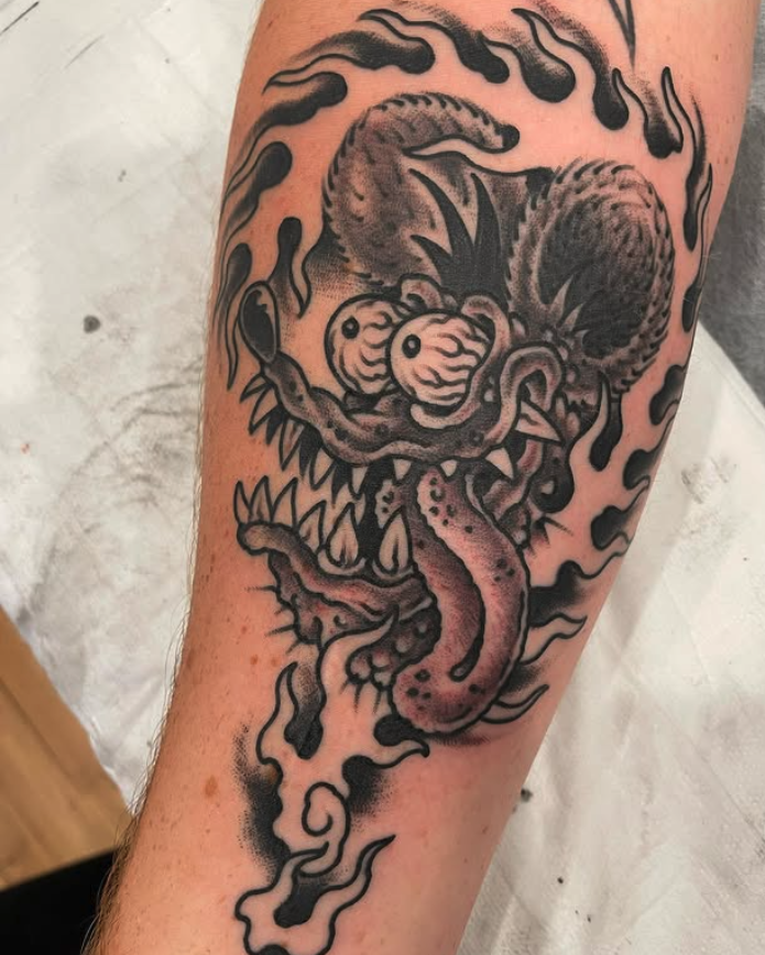 Black and gray traditional-style tattoo of a fierce dragon's face with sharp teeth, extended tongue, and intense eyes, surrounded by swirling flames or smoke.