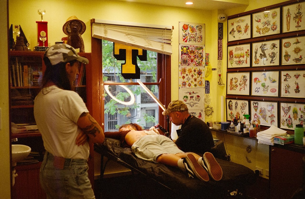 A tattoo artist is tattooing a woman lying on a tattoo bed while another person observes. The room has tattoo designs on the wall and a large window with a yellow letter T decoration.