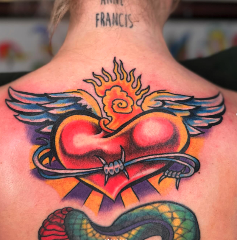 Colorful tattoo of a heart with wings, a sun with a spiral in the center, and a snake below on a person's upper back.