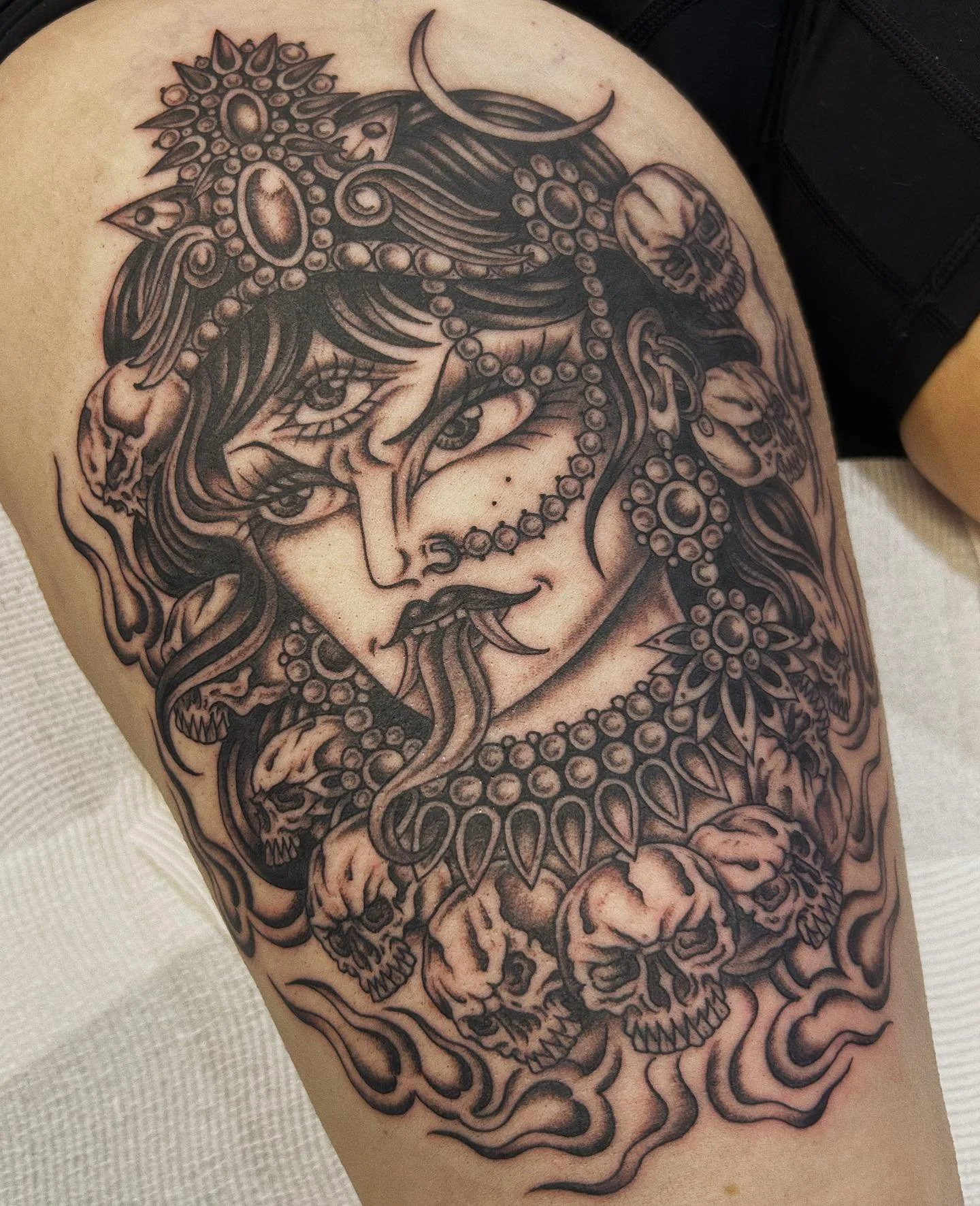 Tattoo of a woman's face with dark hair, wearing jewelry and surrounded by skulls and swirling patterns.