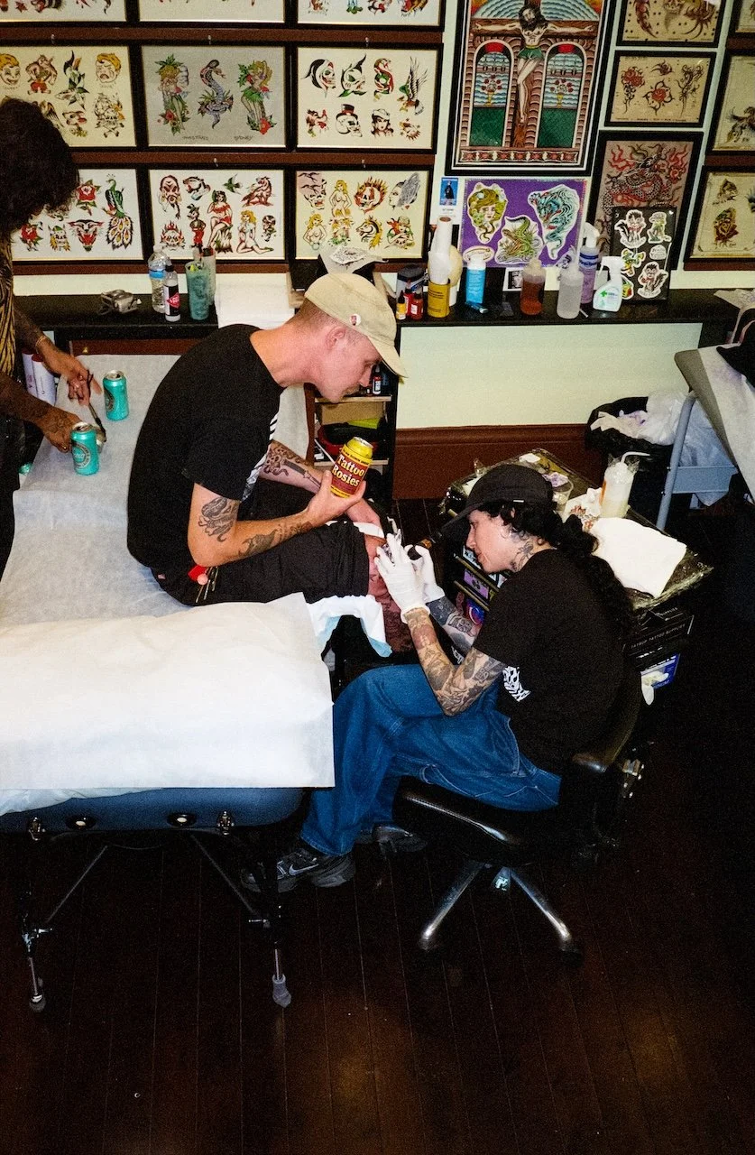 A tattoo artist tattooing a person's thigh while the person sits on a tattoo table. The artist wears a black shirt and has tattoos on her arms. The person receiving the tattoo wears a beige cap and a black shirt. The background features walls with fr