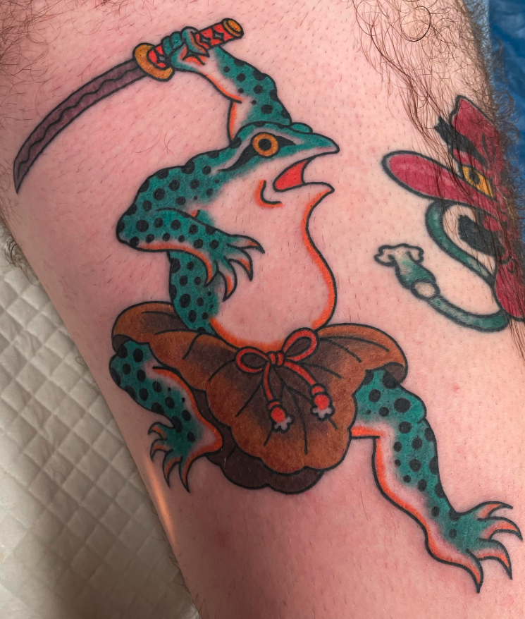 Tattoo of a blue and black spotted gecko with an orange outline, holding a brown butterfly with a red bow on its wings on a person's skin.