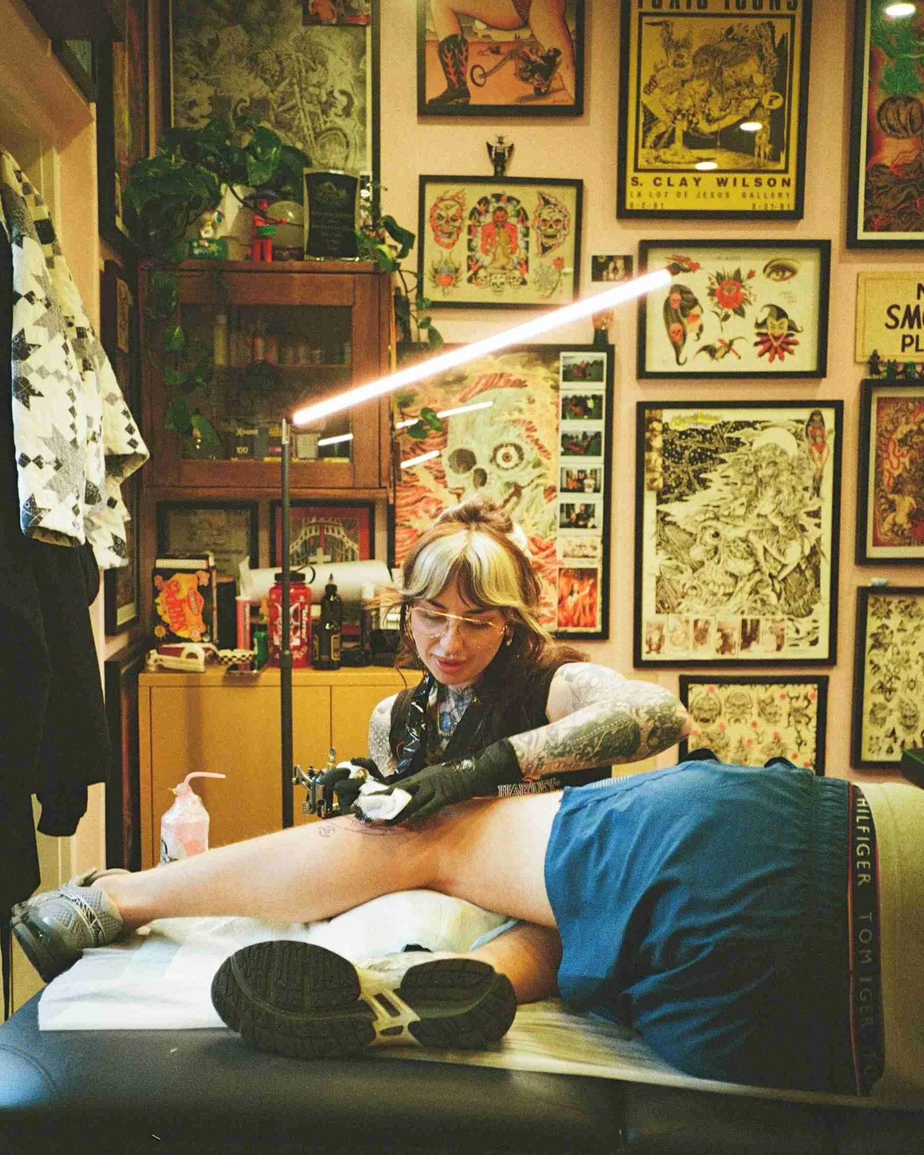 A tattoo artist working on a person's leg in a tattoo studio decorated with various posters and artwork.