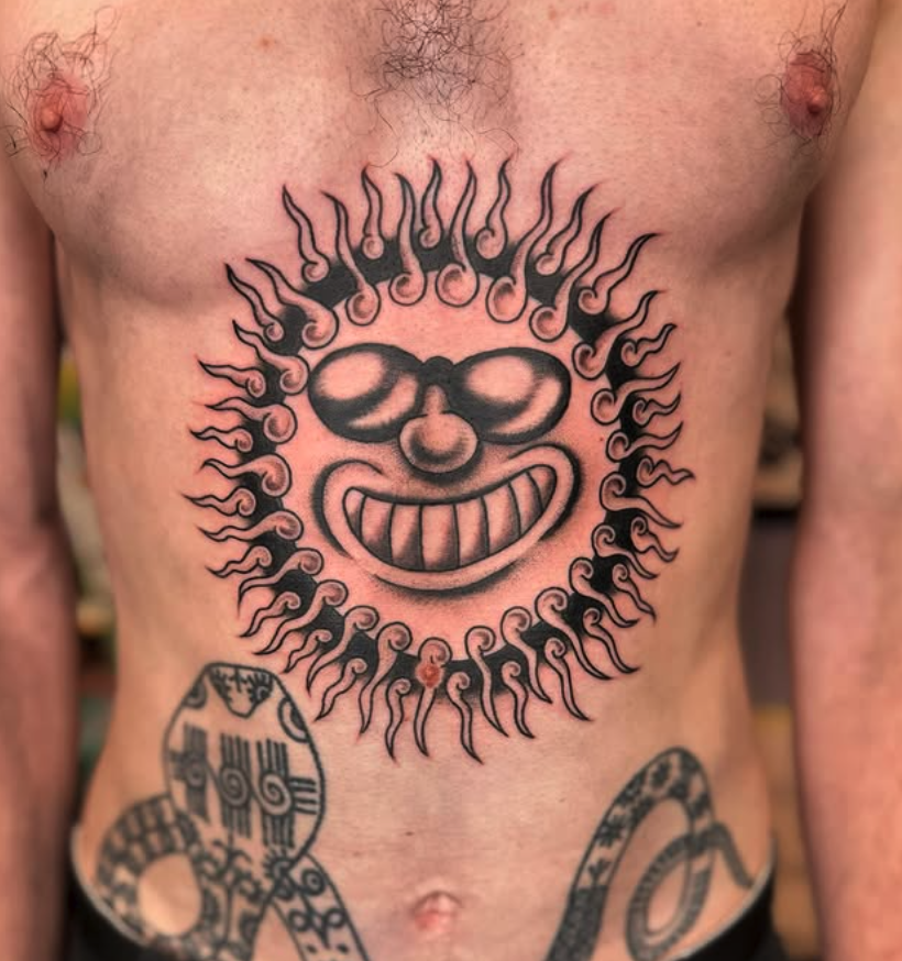 A tattoo of a wide-mouthed, smiling cartoon face with sunglasses, surrounded by stylized flames on a person's chest.