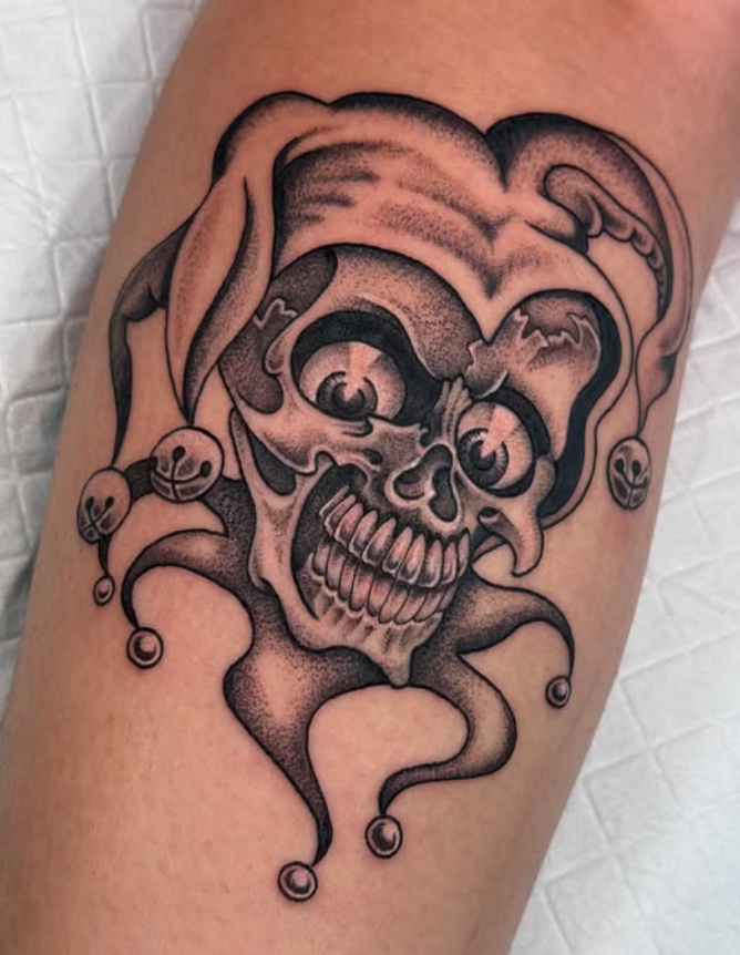 Tattoo of a stylized skull with jester elements, including bells and a jester hat, in black and gray ink.