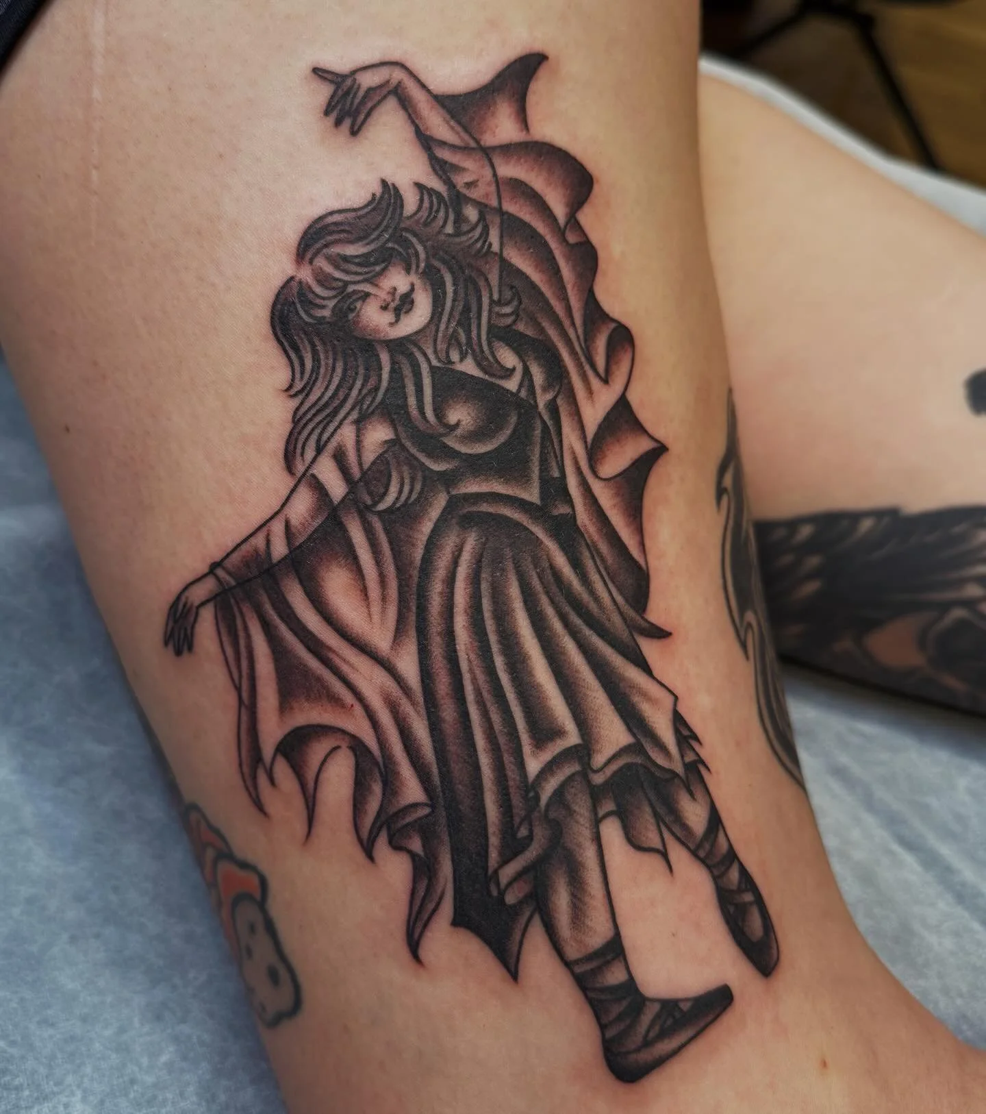 Black and gray tattoo of a fairy with long flowing hair, wearing a dress, with bat-like wings, holding a wand, on a person's arm.