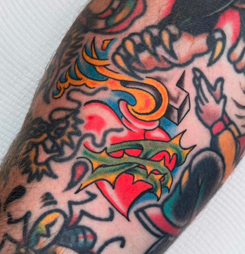 Close-up of colorful tattoo on skin, featuring a red heart, a green dragon, a blue and yellow design, and other cartoon-style illustrations.