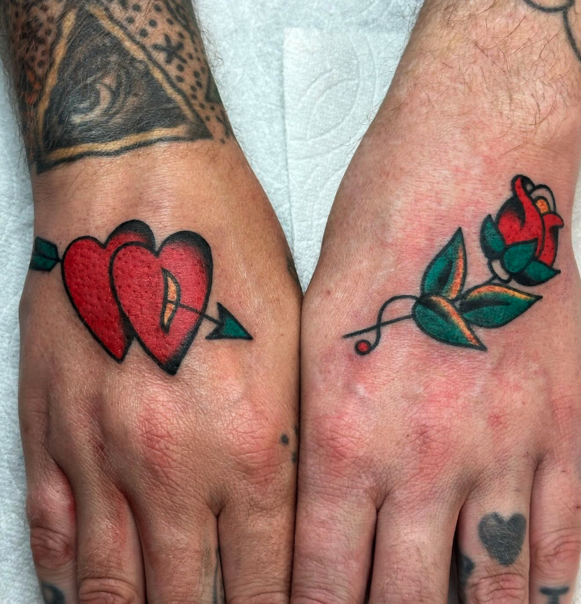 Two hands with tattoos, one featuring two overlapping red hearts with an arrow passing through them, and the other a red rose with green leaves.