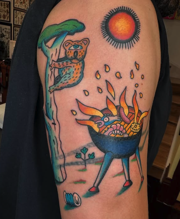 Colorful tattoo featuring a green tree, a leopard climbing the tree, a red and black sun, and a blue bowl of flames with whimsical shapes inside on a person's upper arm.