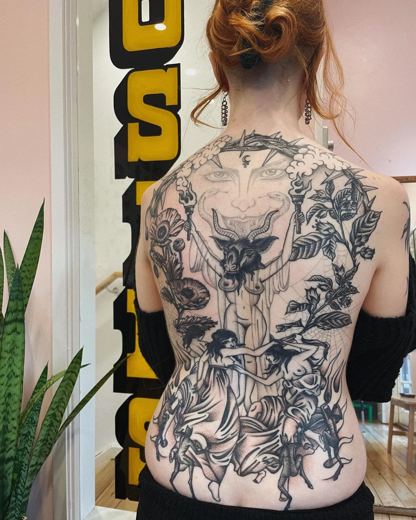 A woman with extensive black tattoo art covering her back, depicting surreal and mythological figures, including a goat-headed creature, women, and a face with multiple eyes, with a woman with wings around her on a tree.