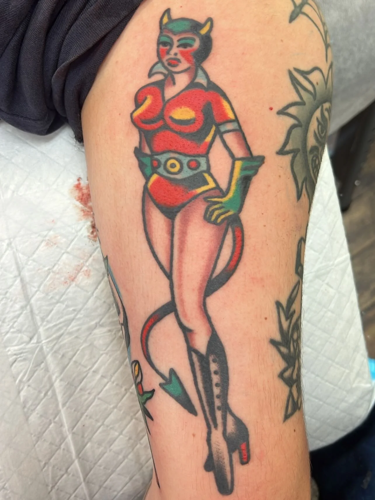 Tattoo of a stylized female devil with horns, red and black outfit, green gloves, high heels, and a tail, standing with one hand on her hip.