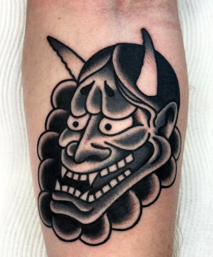 Tattoo of a clown face with a horn, wide smile, and exaggerated features, surrounded by dark shading and cloud-like shapes.