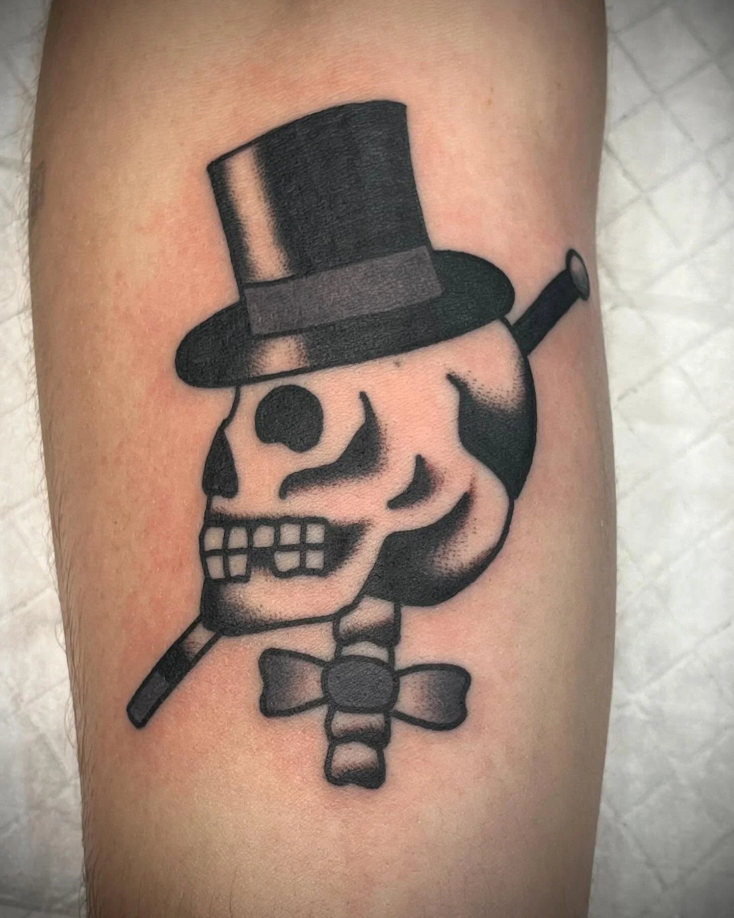 Tattoo of a skull wearing a top hat, with a cigar in its mouth, and a bow tie, all done in black and gray ink.
