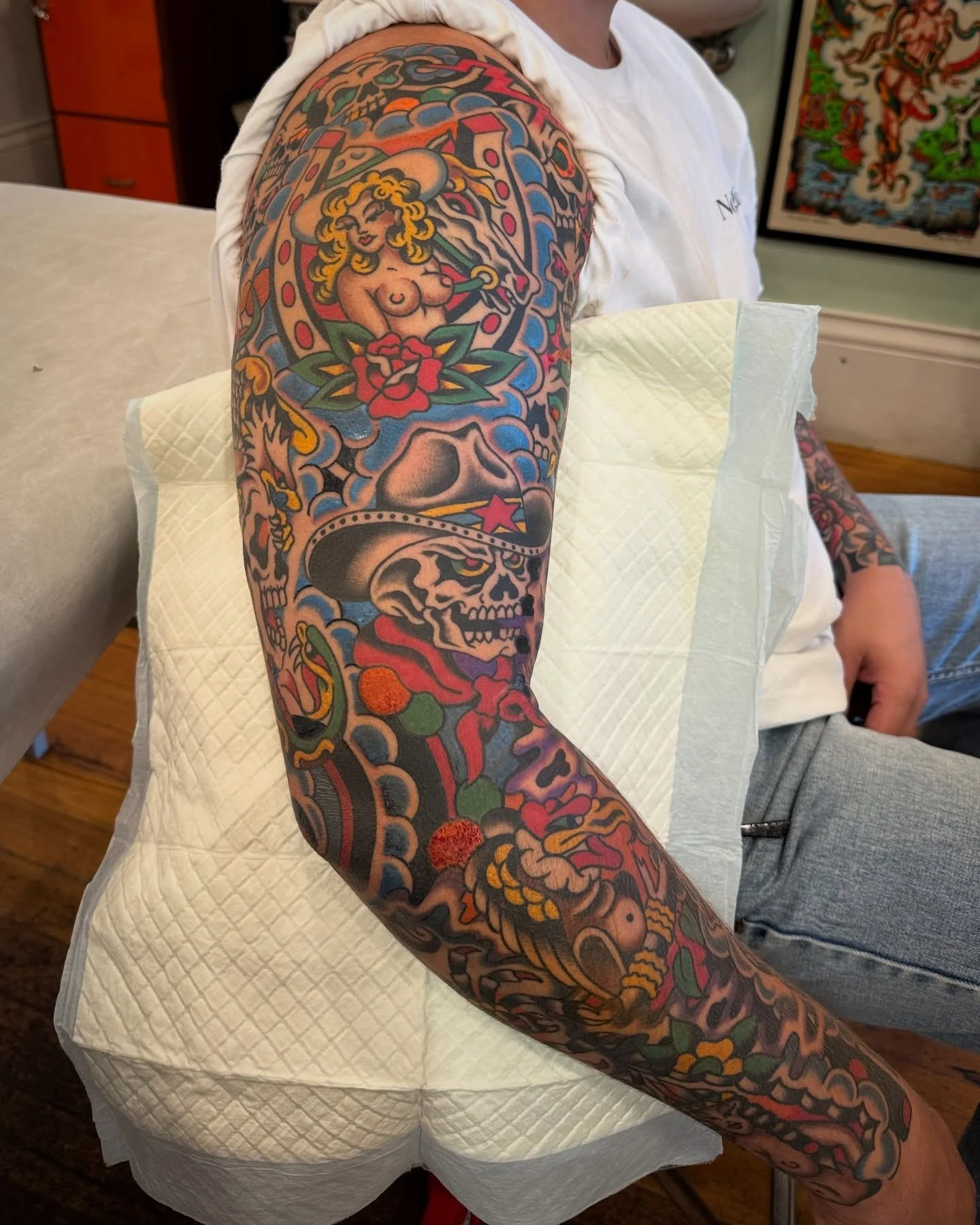 A person's arm with a colorful traditional style tattoo sleeve featuring a woman, a skull wearing a cowboy hat, flowers, and various decorative elements.
