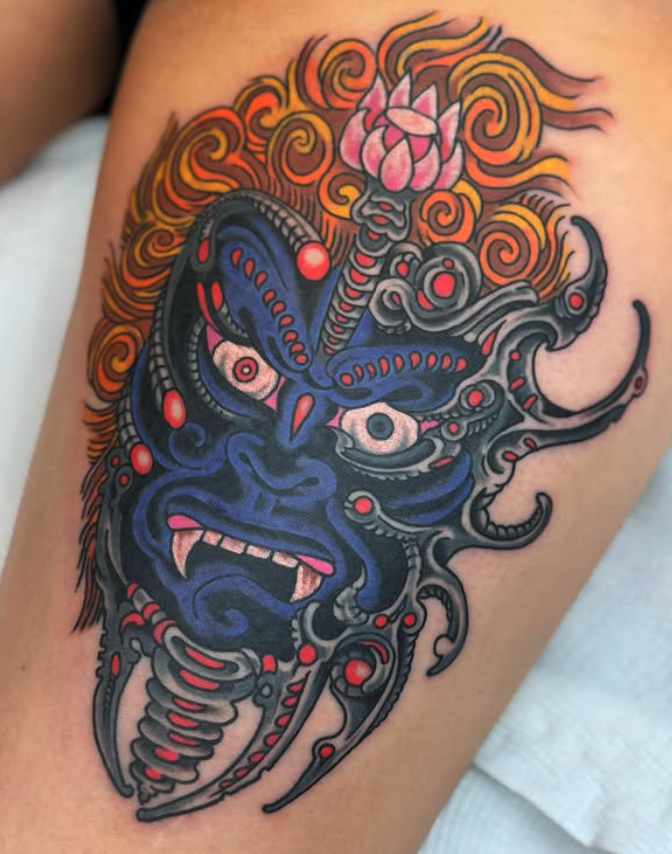 Tattoo of a mythical creature with a demon face, featuring blue and gray skin, bloodshot eyes, and large teeth. The creature has multiple tentacle-like appendages and fiery orange flames surrounding its head, with a pink lotus flower on top.