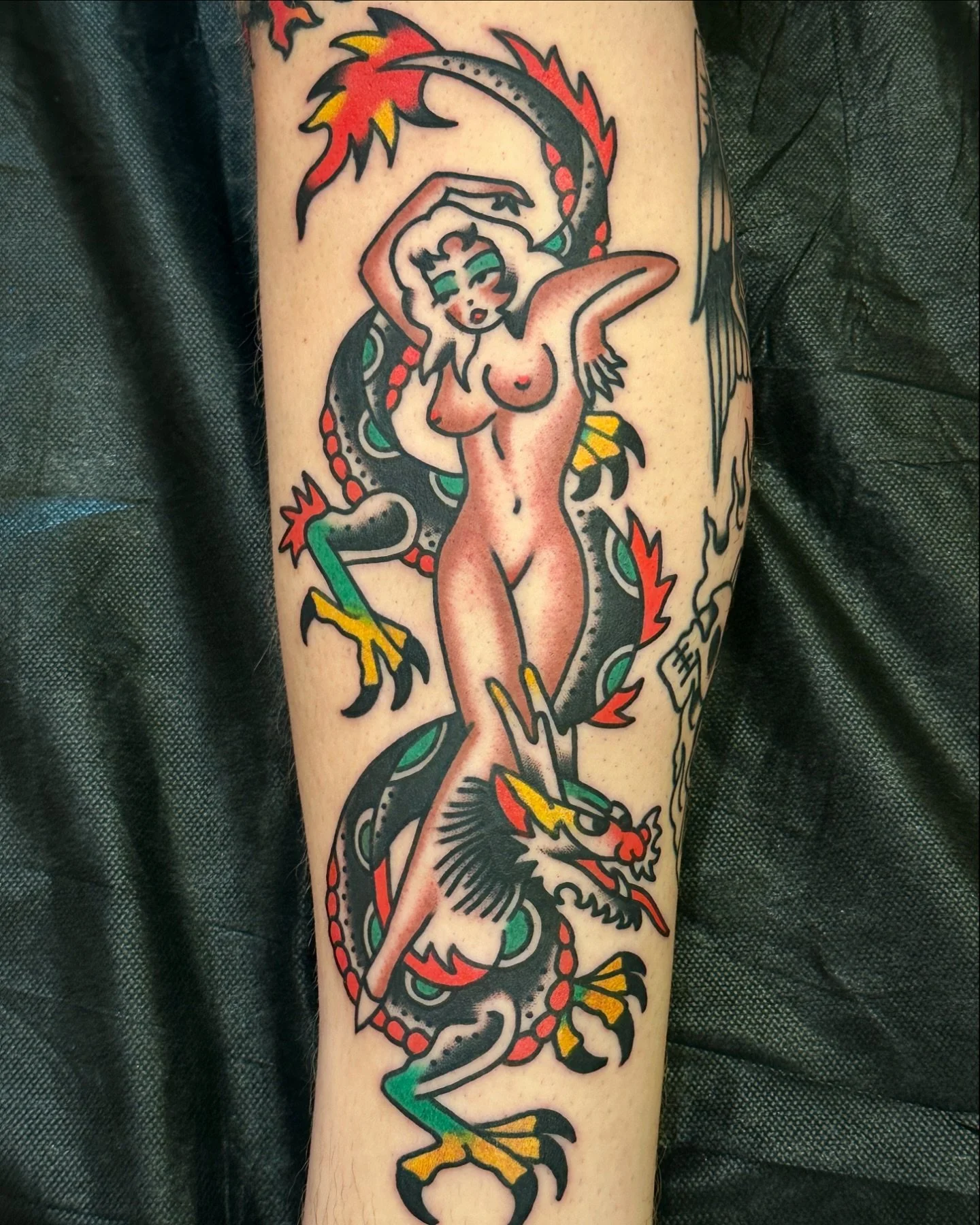 Tattoo of a nude woman with green eyes and dark hair, intertwined with a black dragon with red and green details, surrounded by red and yellow flames.