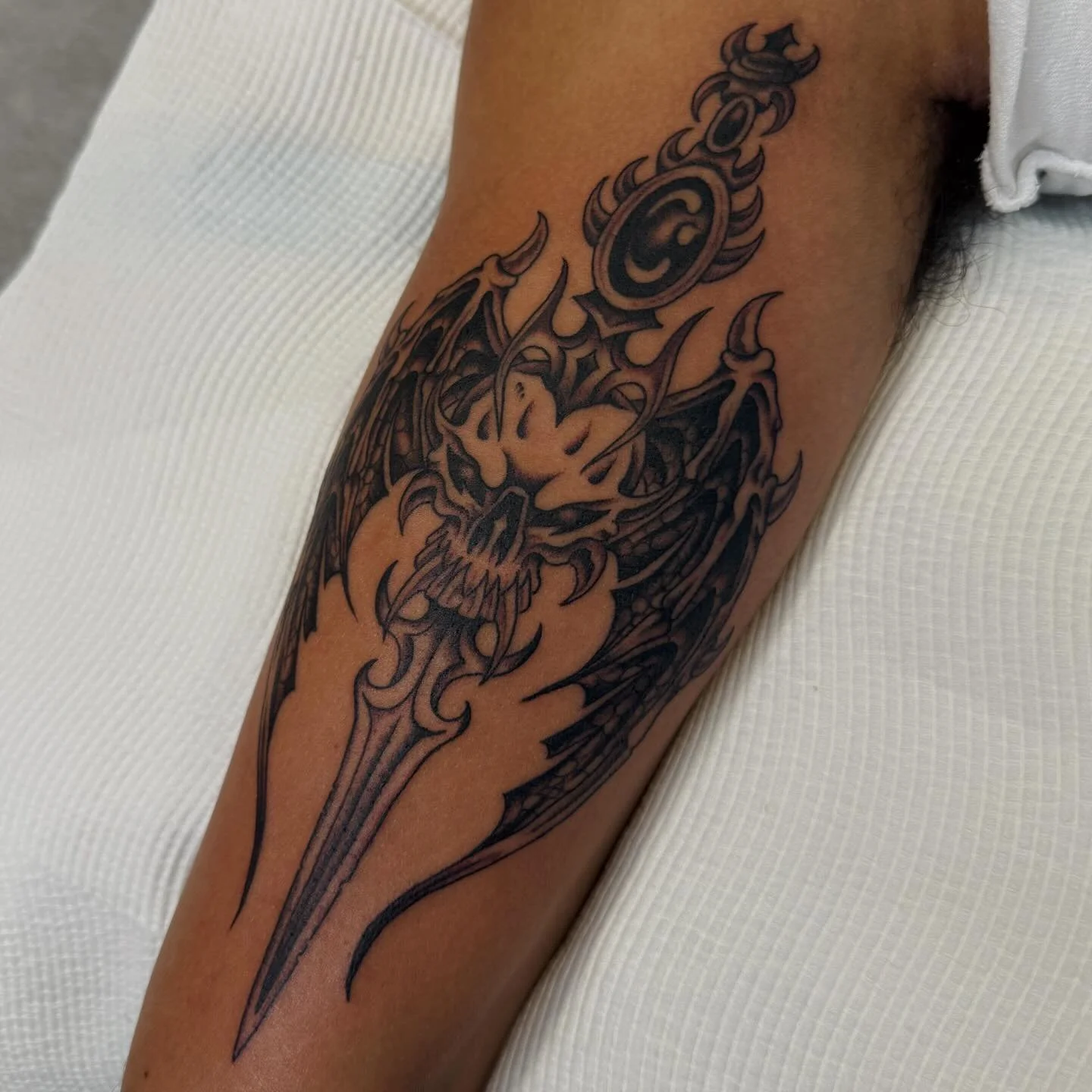 Tattoo of a fierce dragon with a skull face, claws, and a sword, in black ink on a person's arm.