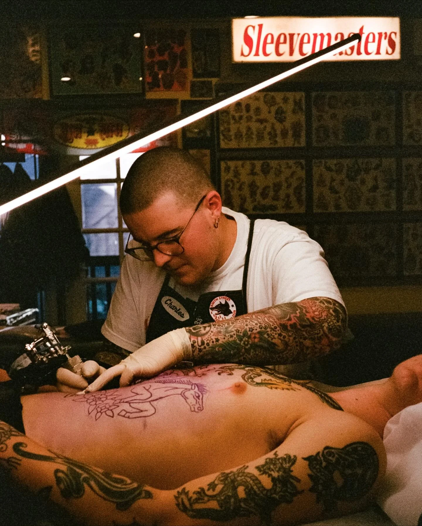 Tattoo artist in black apron working on a person's stomach area in a tattoo shop decorated with posters and a neon sign.