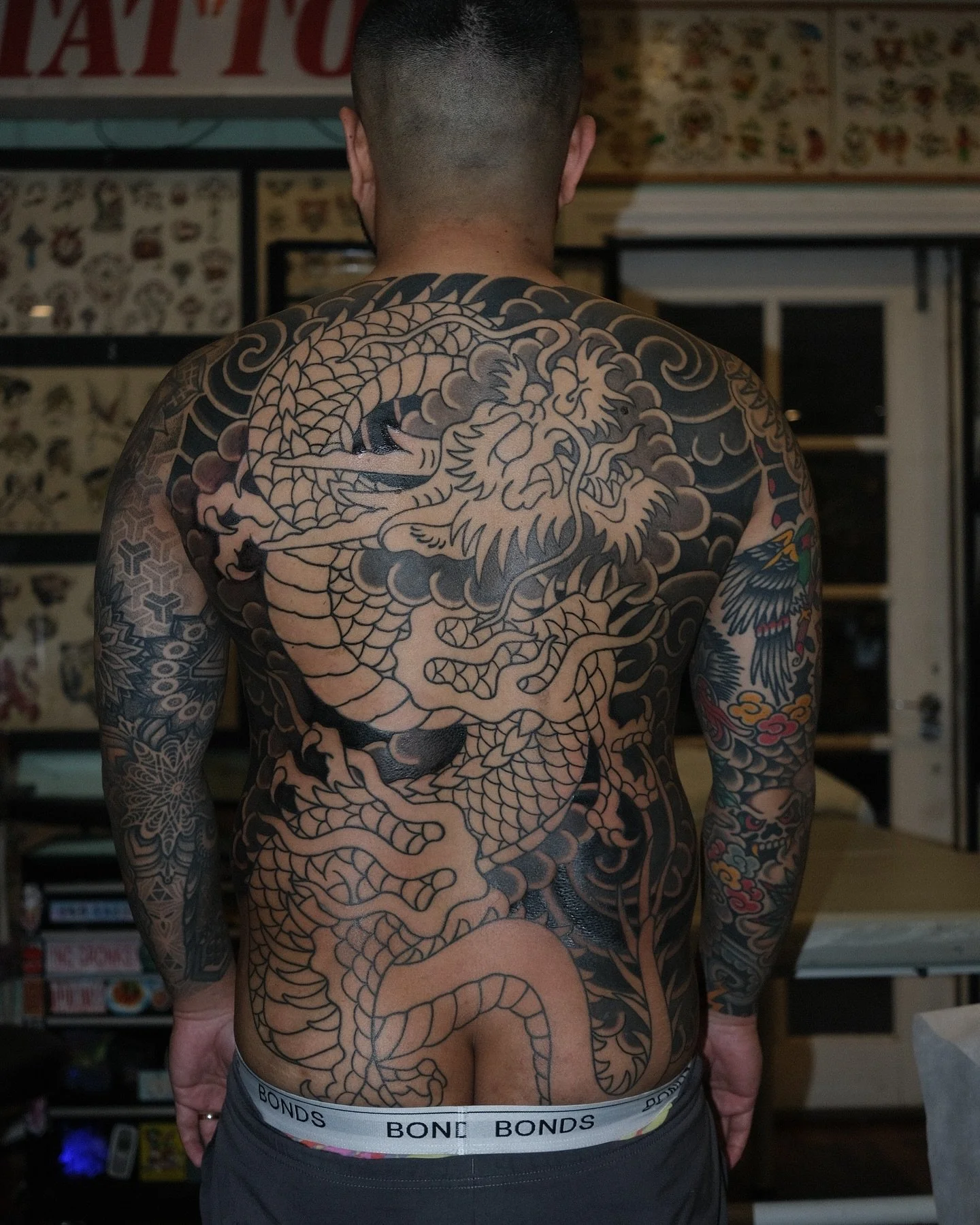 A man with a large, intricate black and gray dragon tattoo covering his back, with additional tattoos on his arms, in a room with tattoo art and posters on the walls.