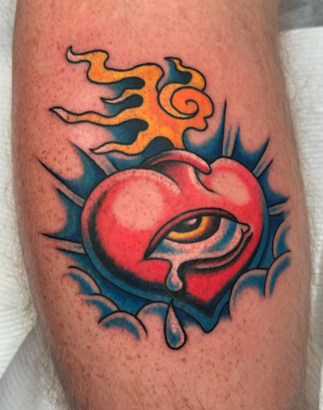 Tattoo of a bleeding, stylized red heart with a yellow eye inside, surrounded by blue clouds and orange flames.