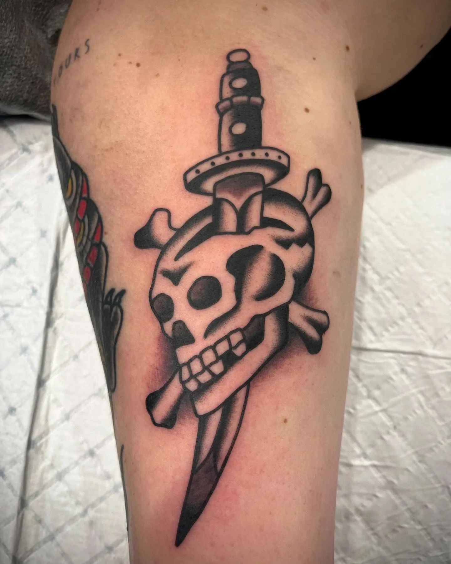 Tattoo of a skull with a sword through it, featuring a face with hollow eyes and nose, and teeth showing, in black and gray ink.