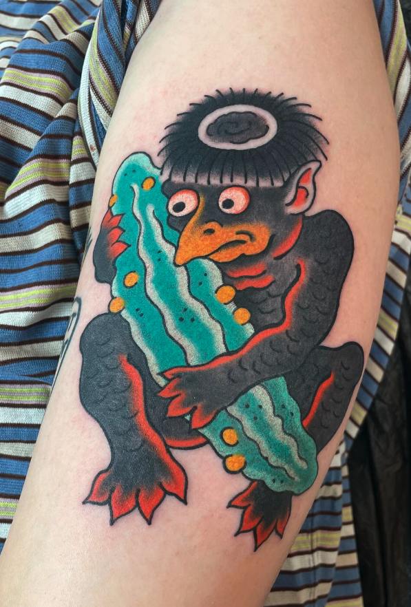 A colorful tattoo of a creature with a bird-like beak, multiple eyes, and a dark, furry head, holding a blue and green fish with yellow spots.