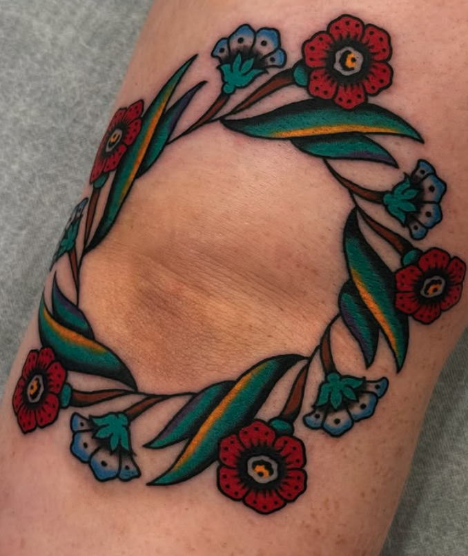 A colorful tattoo of a floral wreath with red flowers, blue flowers, green leaves, and brown branches, arranged in a circular pattern on skin.