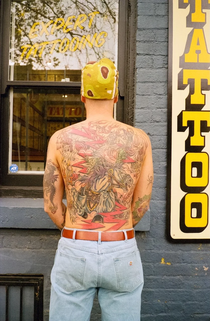 A shirtless person with tattoos on their back and arms, wearing a yellow camouflage hat, facing a tattoo shop window with a sign that reads 'Expert Tattooing'.