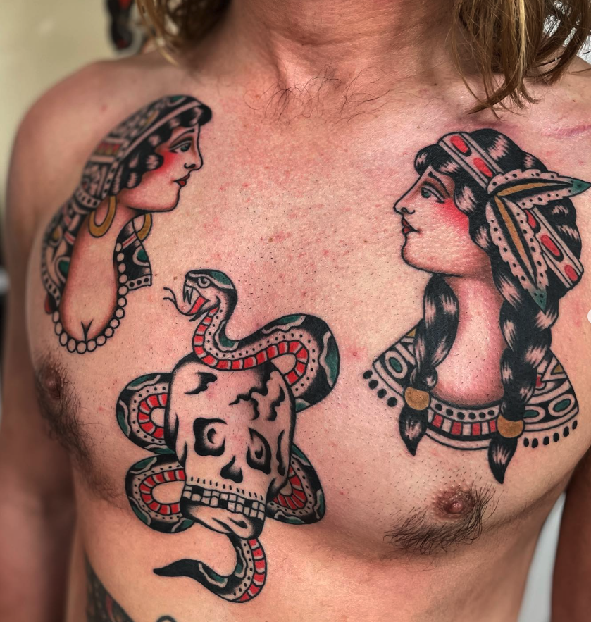 Tattoo of two women with traditional hair, jewelry, and headbands, facing each other. Between them, there is a snake coiled around a skull, with the snake's head emerging above the skull's top.