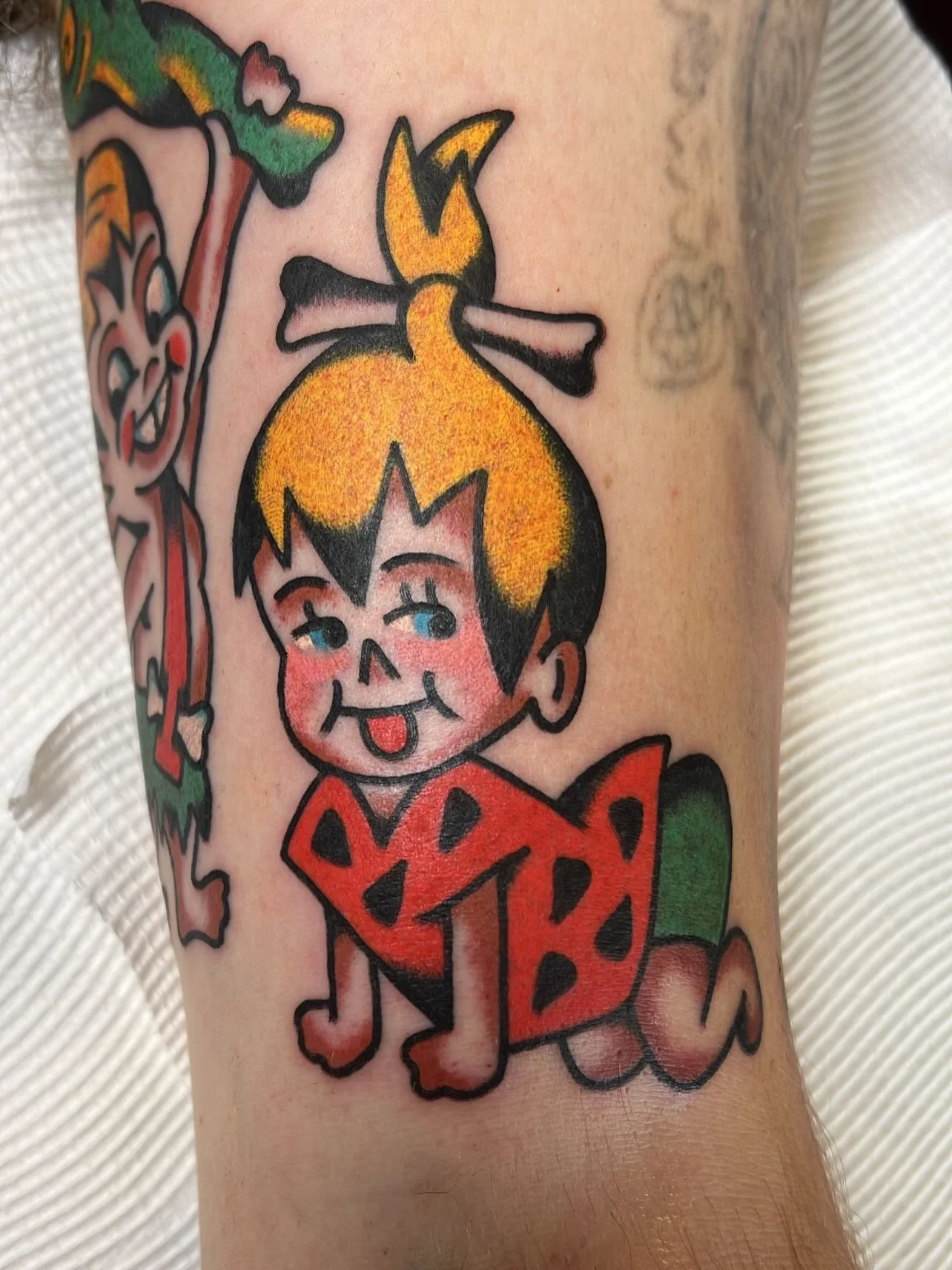 Tattoo of a cartoon boy with yellow hair, sticking out his tongue, dressed in a red outfit with black spots, and a green tail, on a person's arm.