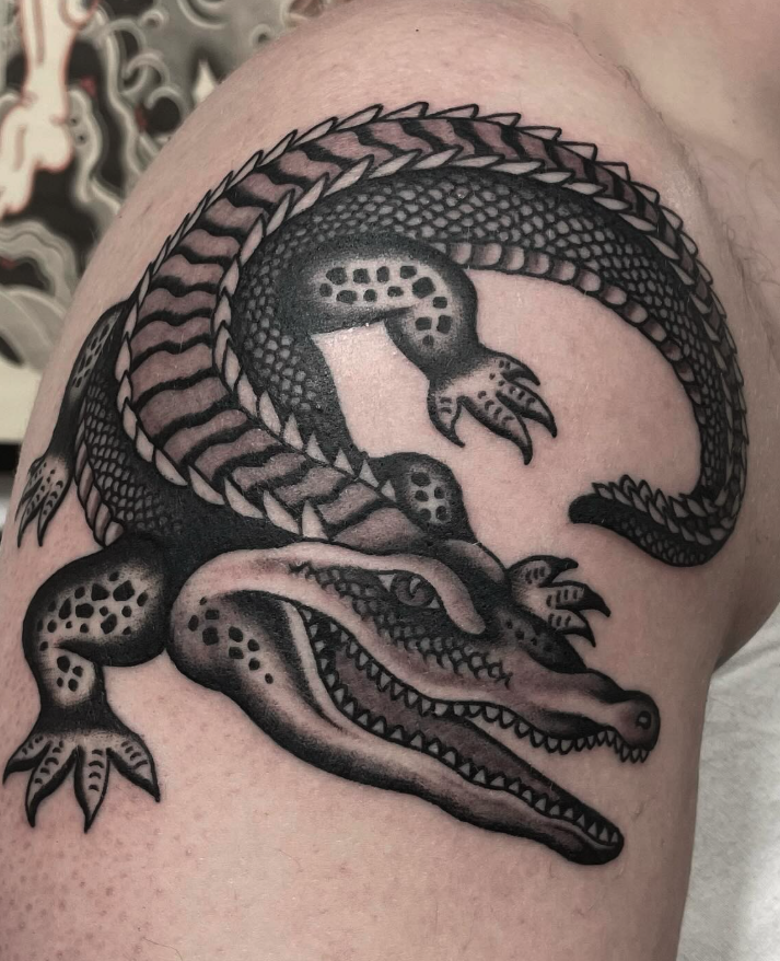 Black and gray tattoo of a crocodile's head and tail on a person's shoulder. The crocodile's head is detailed with sharp teeth and scales, with its body curling around.