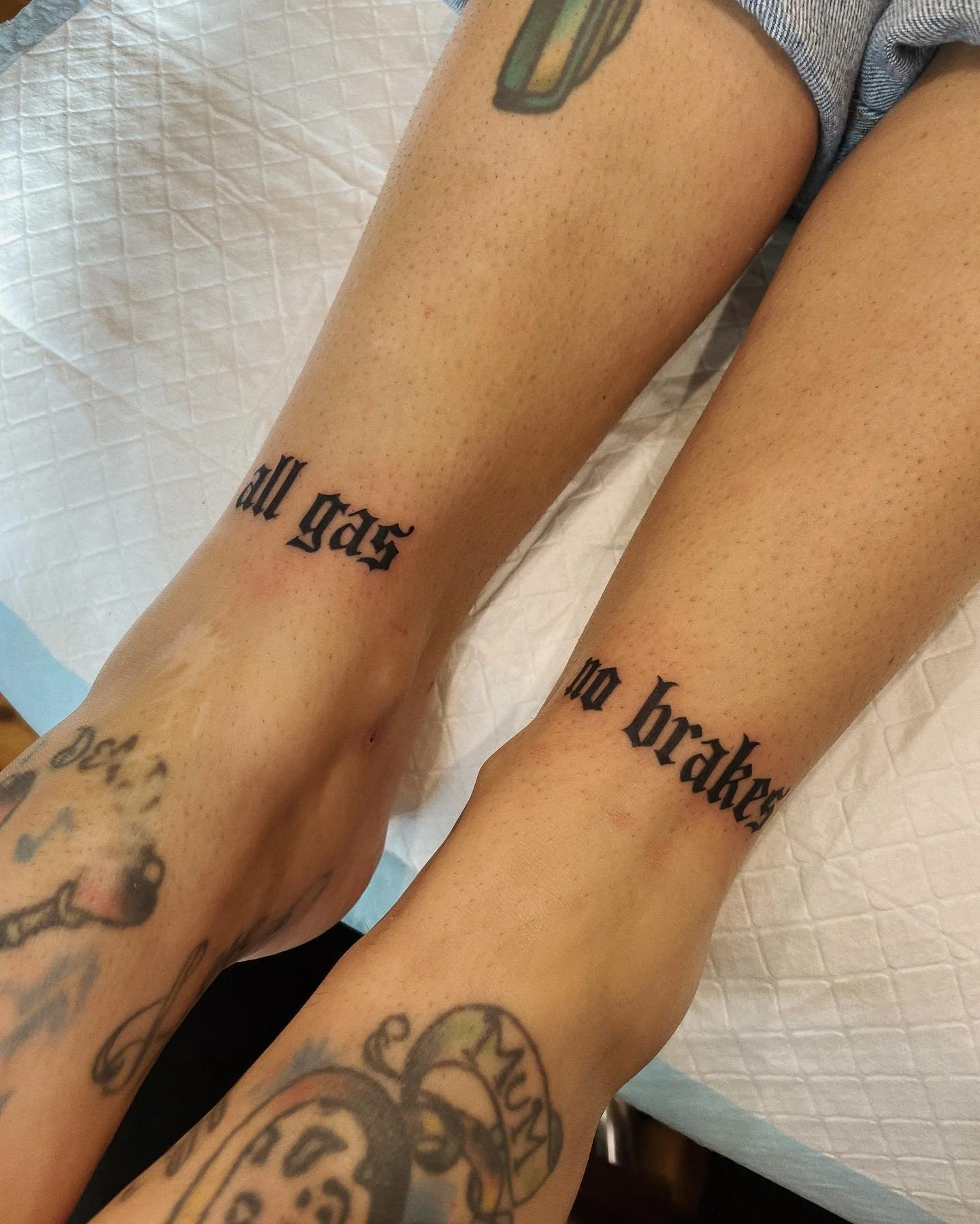 Close-up of two tattooed legs with text tattoos on the shins that read 'all gases' and 'no brakes' in black Gothic font. The person has additional tattoos on their feet and calves.
