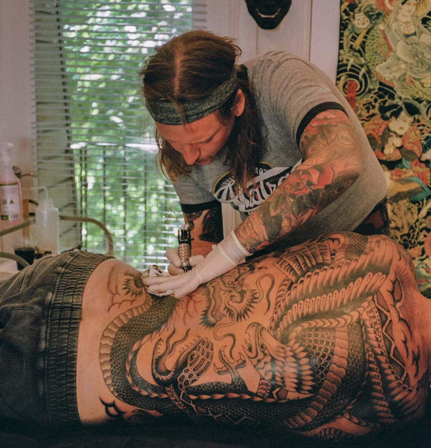 A tattoo artist working on a large tattoo on a person's back, featuring a dragon surrounded by traditional Japanese design elements.