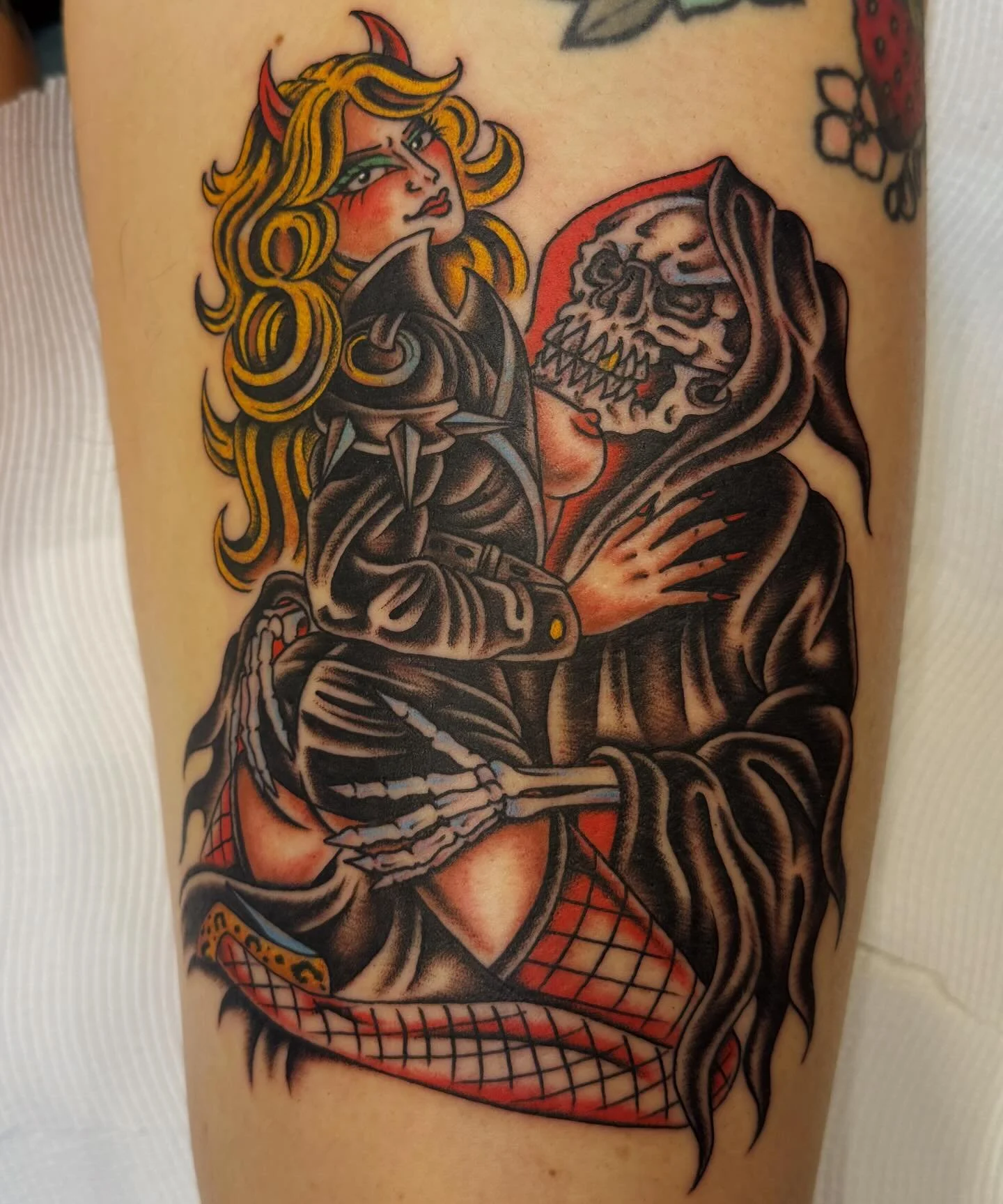 Tattoo of a devil woman with horns, long blonde hair, a black leather jacket, and a fishnet stocking, holding a skeleton with a skull face and a hood, on a person's arm.