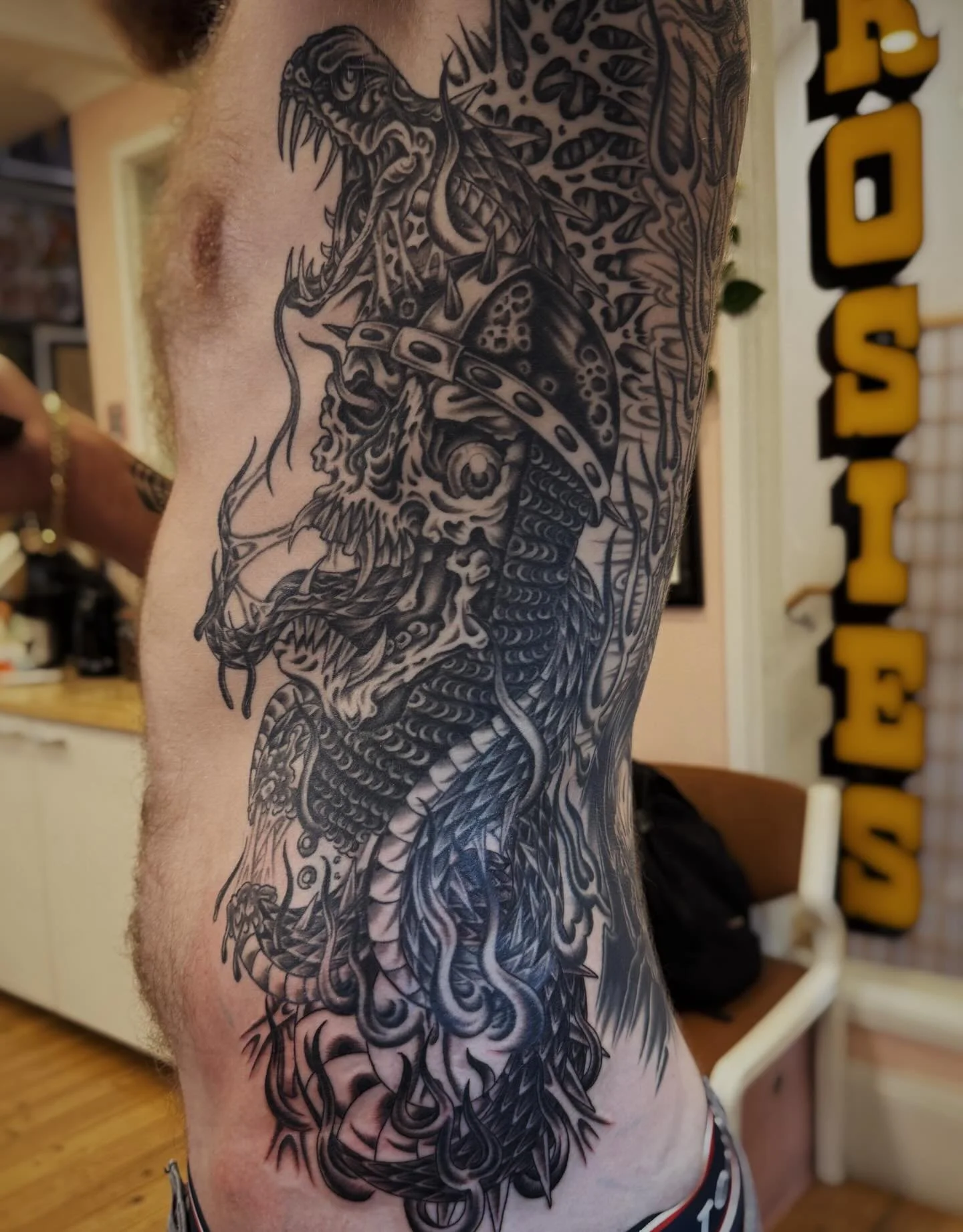 Black and gray tattoo of a fierce, stylized dragon's head and upper body, with intricate details and shading, covering a person's side torso.