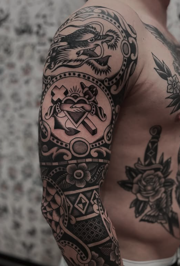 Black and grey tattoo sleeve featuring a dragon's head, a heart with a sword and chain, and intricate geometric and floral patterns.