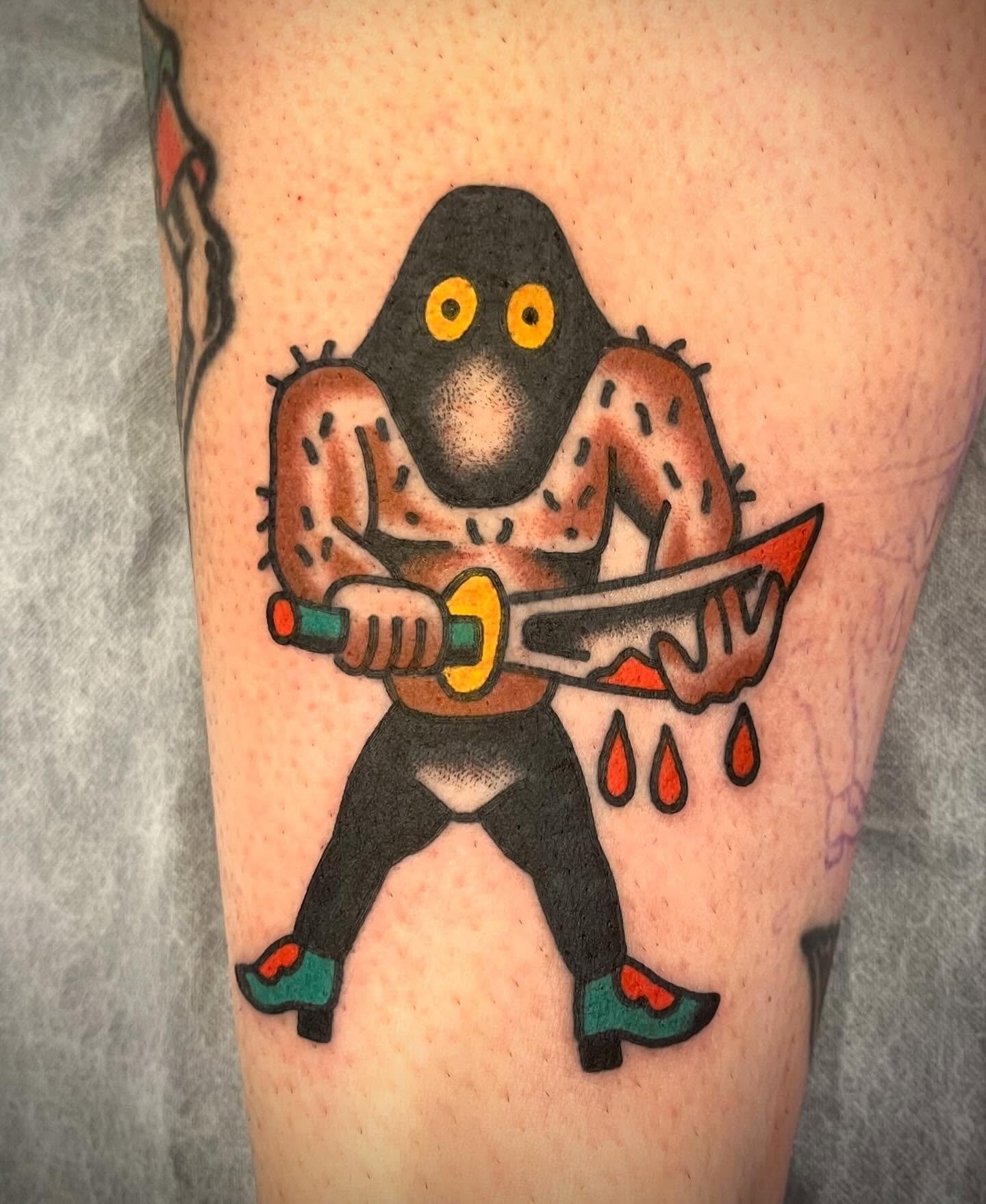 A tattoo of a cartoon character with a black head, yellow eyes, and a brown muscular body holding a bloody sword.