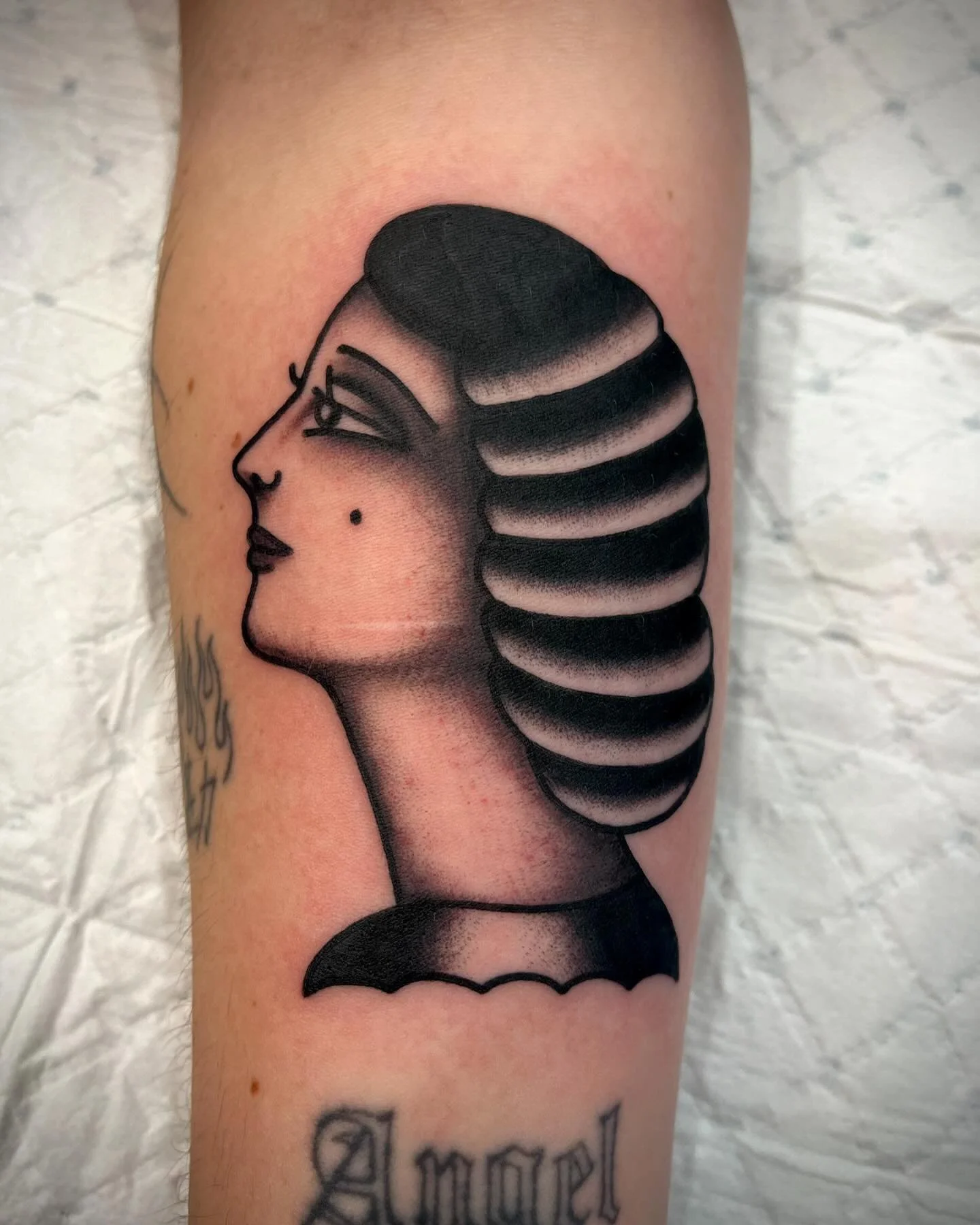 Tattoo of a stylized woman’s profile with black hair in banded style, detailed face with closed eye, lips, and a mole. The tattoo incorporates shading for depth and detail.
