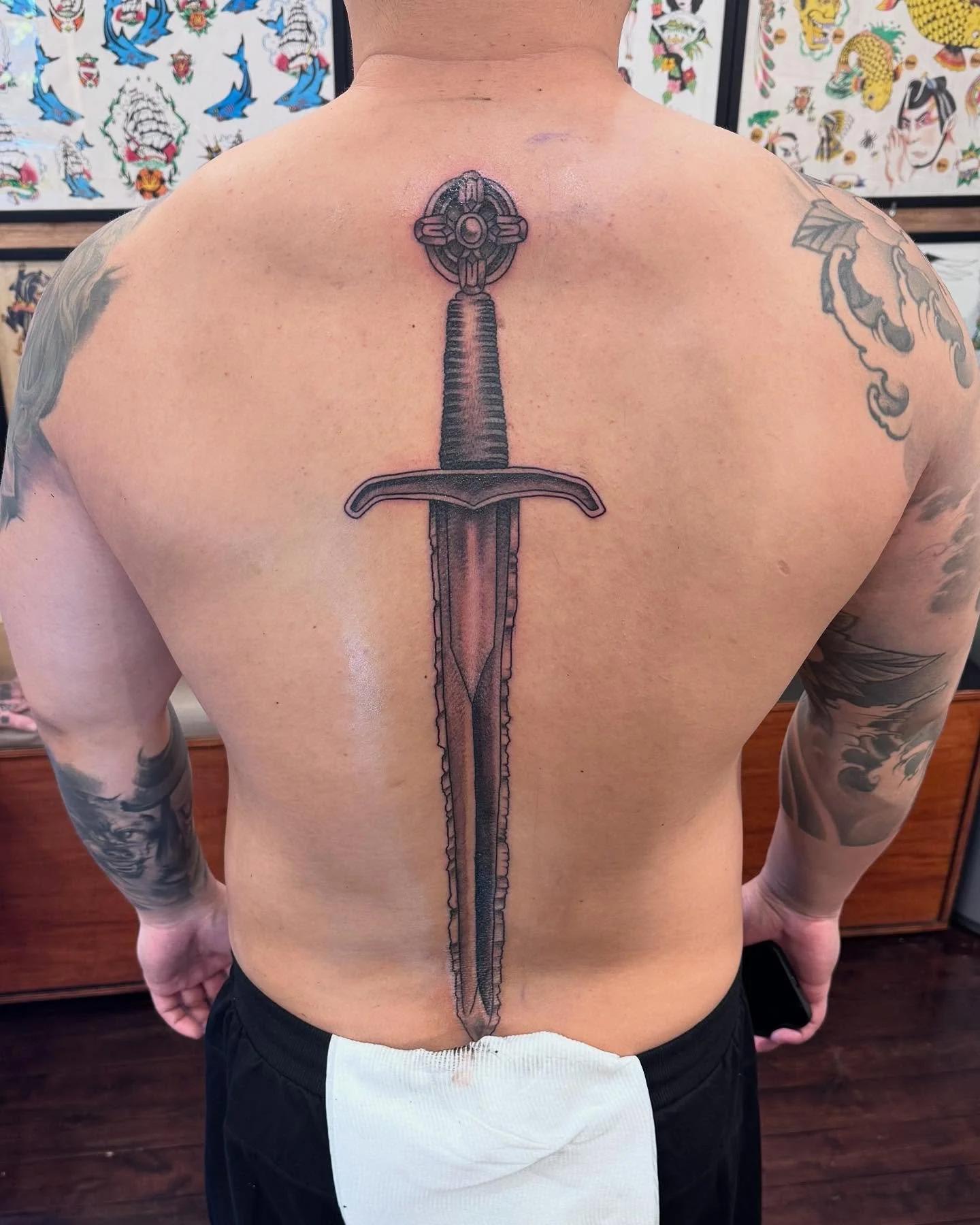 Tattoo of a detailed sword on a person's back, with a decorative hilt at the top and a long, straight blade extending downward.