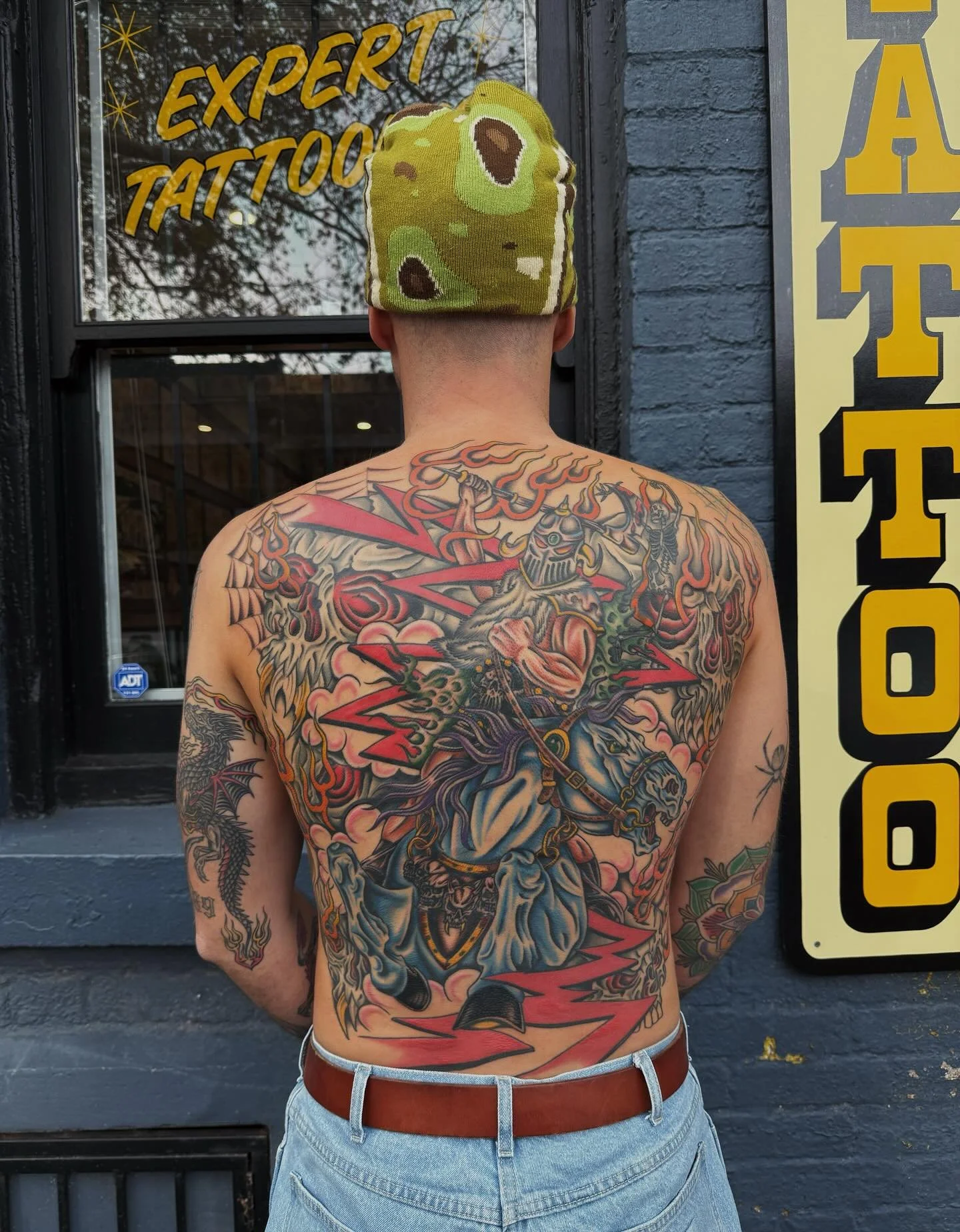 A man with a large, detailed tattoo covering his back, featuring a knight on a horse, surrounded by flames and lightning, standing in front of a dark brick wall; he is wearing a green camouflage beanie and light blue jeans with a brown belt.