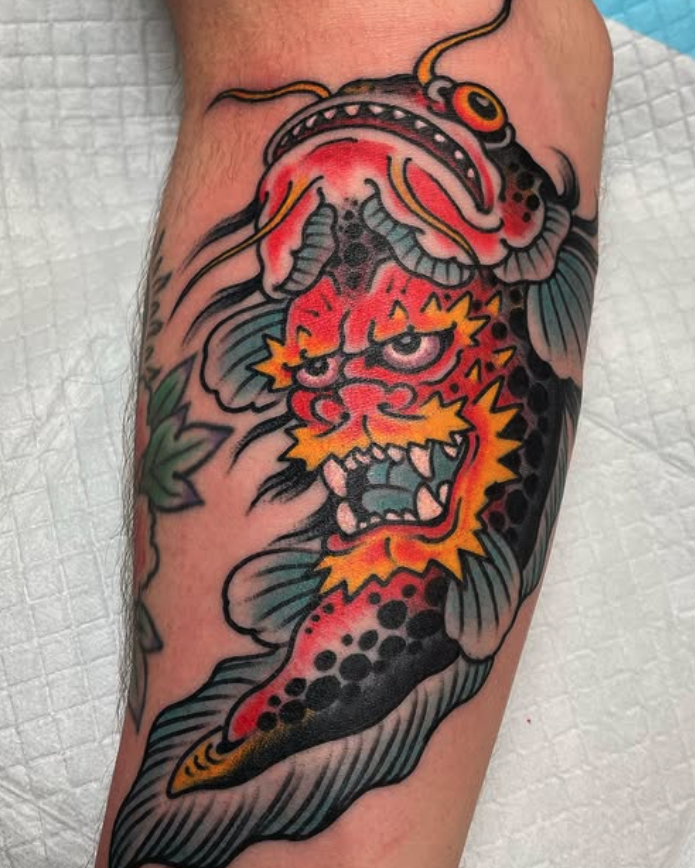 Tattoo of a fierce tiger with a fish on its head, featuring bold black outlines, red and orange coloring, and green leaves in the background.