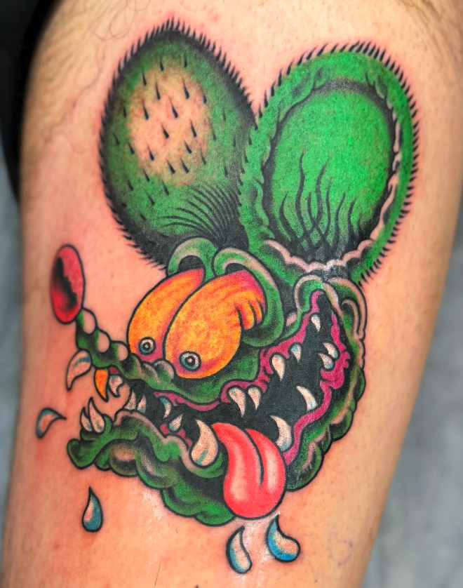 Tattoo of a cartoonish green creature with large ears, sharp teeth, and a long tongue, featuring exaggerated facial features and bright colors.