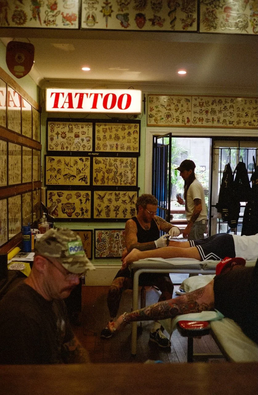 Tattoo shop with clients getting tattoos and tattoo artists working, decorated with tattoo flash art on the walls, and a bright illuminated tattoo sign.