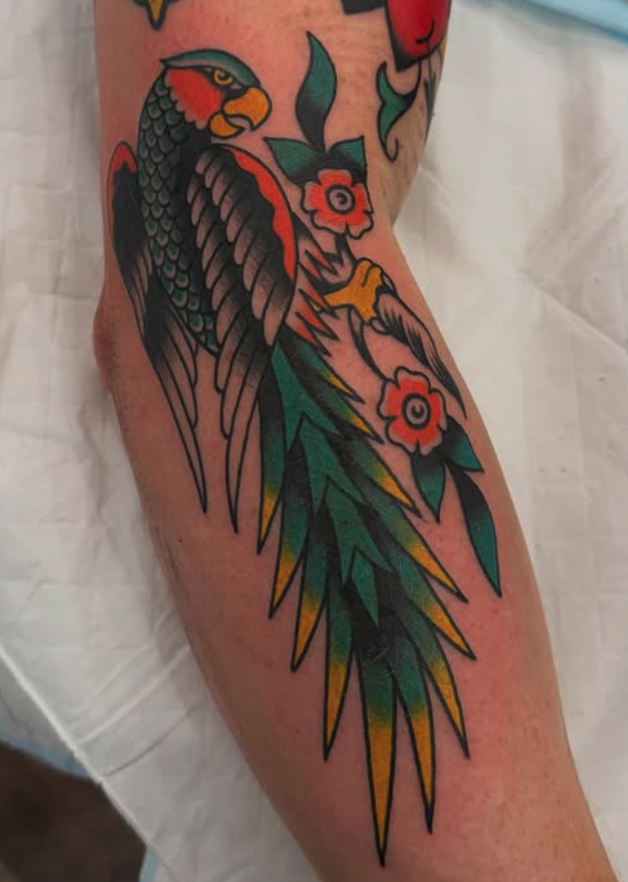 Tattoo of a detailed, colorful bird resembling a phoenix or hawk, with orange and yellow accents, perched among pink flowers with green leaves.